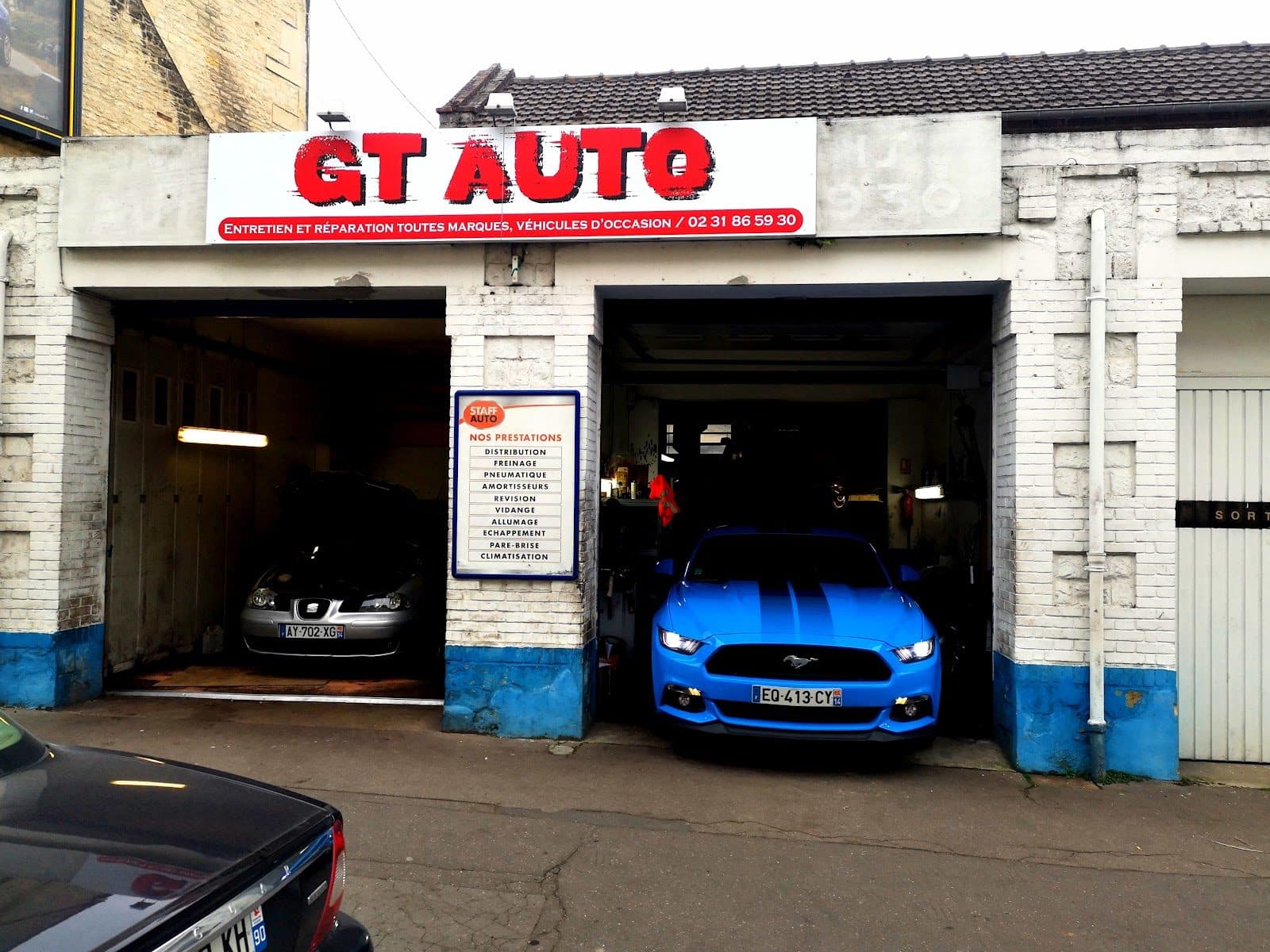 Les deux baies de GT AUTO ouvertes : une Seat capot levé à gauche, une Ford Mustang bleue à droite, et entre les deux le panneau « Nos prestations » vissé au pilier.