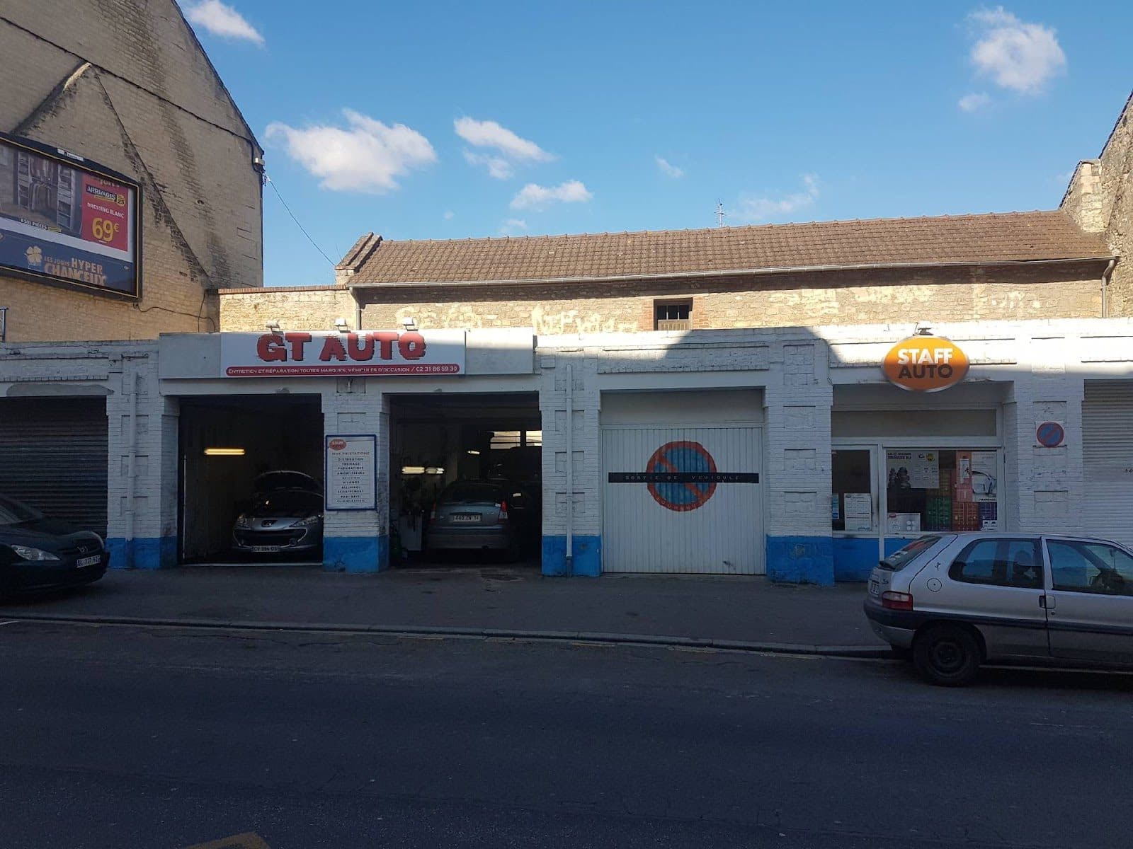 La façade de GT AUTO en plein soleil, les deux baies ouvertes avec des véhicules à l’intérieur et le panneau des prestations sur le pilier central.