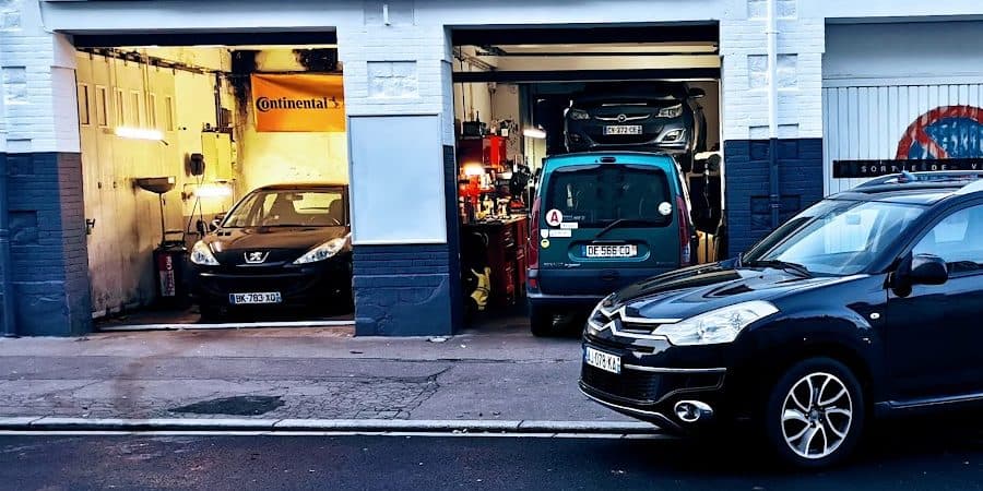 Les deux baies de l’atelier GT AUTO vues depuis la rue : une berline au sol, un utilitaire et une voiture sur le pont, servante à outils et bâche Continental.