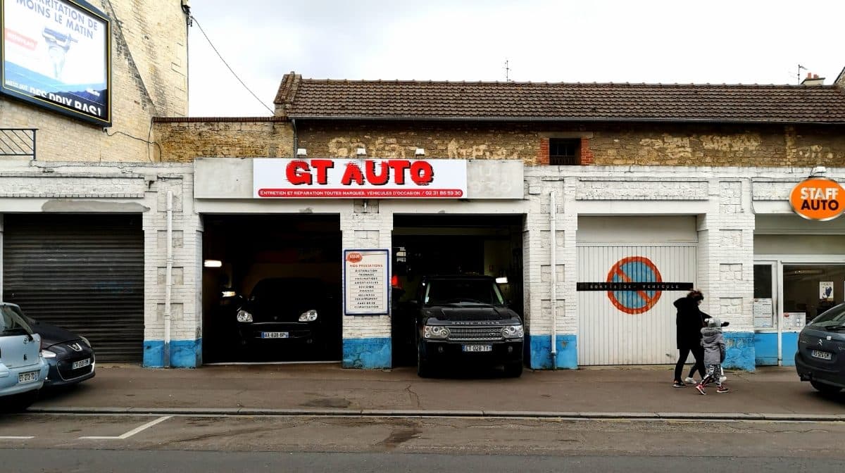 Les baies de GT AUTO ouvertes sur la rue : une Porsche dans la baie de gauche, un Range Rover dans celle de droite, des passants sur le trottoir.