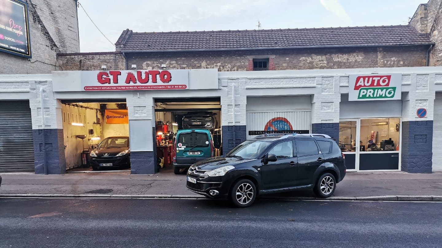 La devanture de GT AUTO, 78 rue de Bayeux à Caen : l’enseigne rouge, deux baies d’atelier ouvertes et éclairées, des véhicules en cours d’intervention.