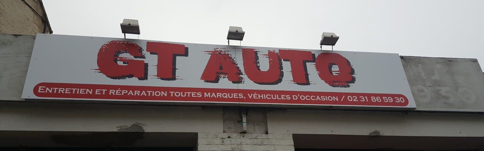 L’enseigne rouge et blanche de GT AUTO vue depuis le trottoir : « GT AUTO — Entretien et réparation toutes marques, véhicules d’occasion / 02 31 86 59 30 ».