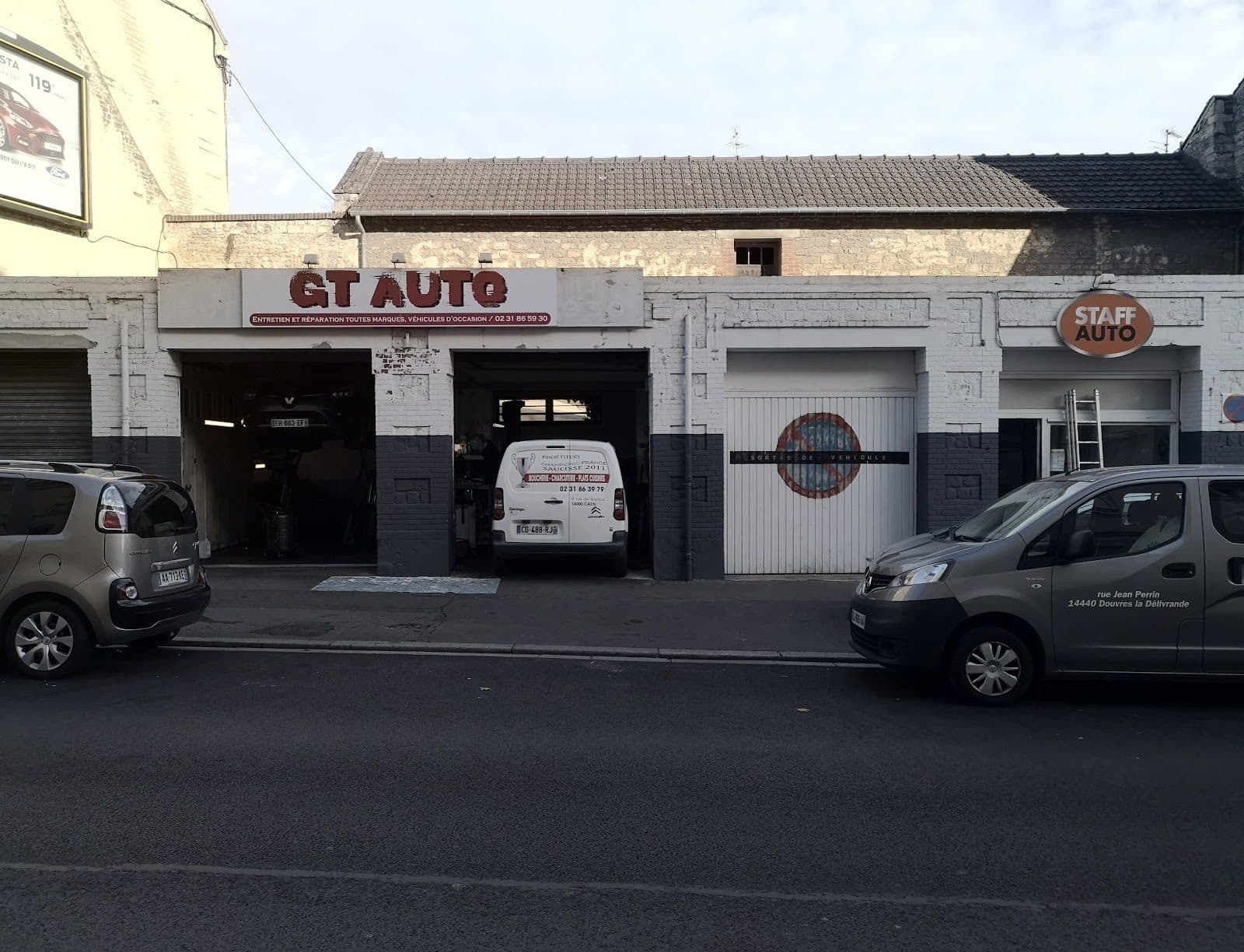 La façade de GT AUTO en fin de journée, baies ouvertes, un utilitaire Berlingo à l’intérieur et des véhicules garés le long du trottoir rue de Bayeux.