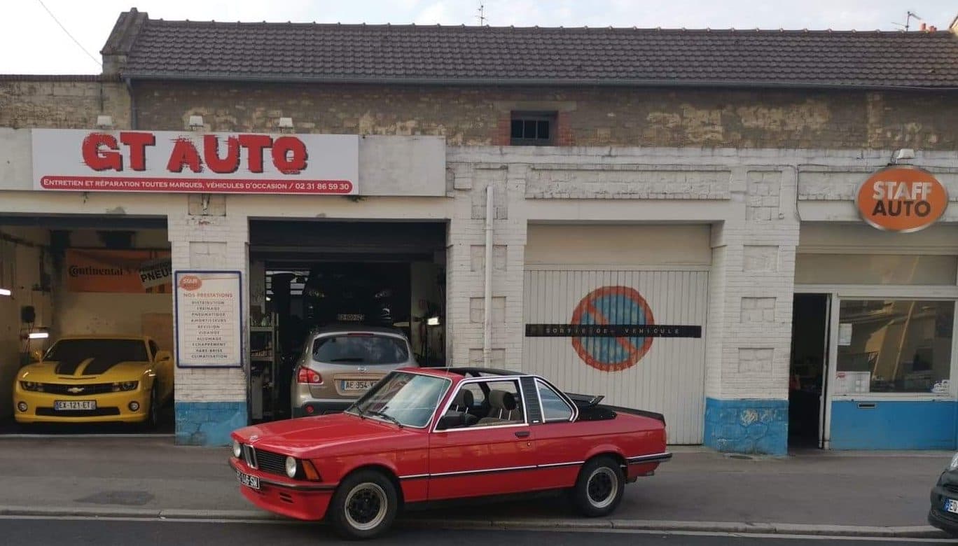 La façade de GT AUTO rue de Bayeux avec un coupé BMW rouge garé devant et une Chevrolet Camaro jaune dans la baie de gauche.